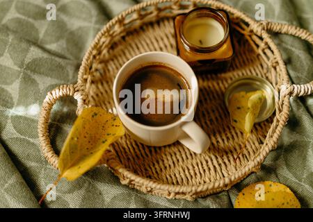 Gemütliche Herbst-Stillleben mit einer Tasse frischem Kaffee, einer warmen Kerze, gefallenen Blättern und einem gewebten Weidenkorb auf texturiertem grünem Stoff bei weichem natürlichem Licht. Stockfoto