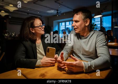 Reife Frau und Mann sitzen an einem Cafétisch und besprechen ein Smartphone während eines abendlichen Gesprächs. Freunde teilen Alltagsmomente in einem gemütlichen Restaurant Stockfoto