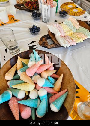 Holzschüssel gefüllt mit einer Vielzahl von farbenfrohen Eiswaffeln, darunter blau, rosa und gelb, auf einem Tisch und Bechern. Obst bei einer Geburtstagsfeier wi Stockfoto