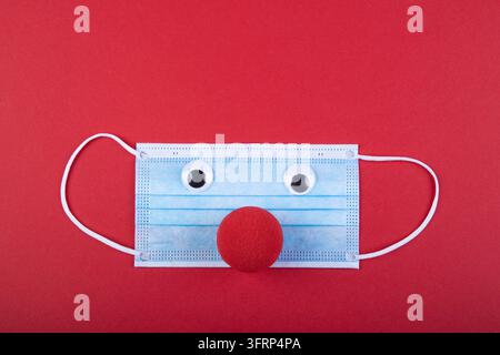 Eine rote Schaumstoffnase liegt unter einer Gesichtsmaske, Symbol für Einschränkungen des Karnevals durch das Coronavirus, roter Hintergrund Stockfoto