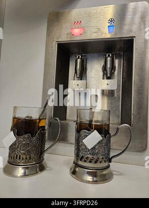 Zwei Tassen Tee stehen auf einem silbernen Tablett neben einem Wasserspender. Der Spender hat einen Knopf, auf dem in Sotschi "Kaffee" steht Stockfoto