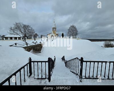 Schneebedeckter Hügel mit einer Kirche und einem Zaun. Der Zaun ist mit Schnee bedeckt. Es gibt Leute, die im Schnee spazieren gehen. Igumenka Stockfoto
