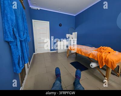 Person sitzt auf dem Boden in einem Raum mit einer blauen Wand. Die Person trägt blaue Pantoffeln. Blauer Raum mit einem hellbraunen Massagetisch. Entspannungsbademantel Stockfoto