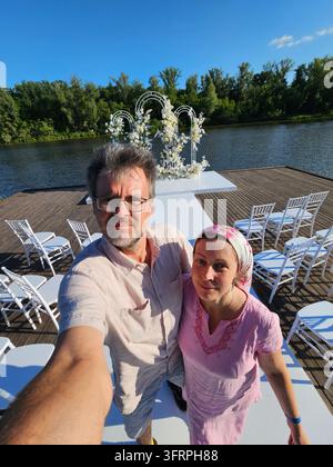 Ein Mann und eine Frau stehen auf einem Holzdeck an einem weißen Hochzeitsblumenbogen. Der Mann macht ein Foto von der Frau Stockfoto