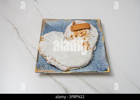 Herzförmiges Palmier-Gebäck mit weißer Glasur, belegt mit Kekskrümeln und einem Keks, serviert auf einem blauen quadratischen Teller. Stockfoto