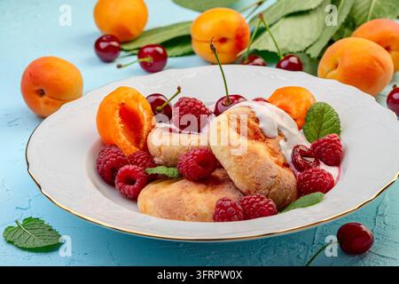 Leckeres Frühstück - Hüttenkäse-Fritters, Hüttenkäse-Pfannkuchen mit Himbeeren, Kirschen, Aprikosen und Rahmsoße auf einem weißen Teller, selektiv Stockfoto
