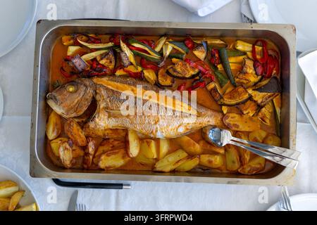 Oberansicht eines Stahltabletts mit einem ganzen goldenen gebackenen Brachsen, der auf Kartoffelkeilen, Zucchini, Aubergine und roten Paprikaschoten in einer orangefarbenen Kochsauce liegt, wi Stockfoto
