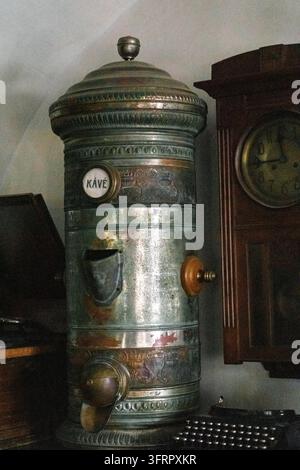 Ein antiker Metallkaffeemühle steht neben einer verzierten Holzuhr in einem Vintage-Cafe-Interieur. Die Mühle trägt das Wort „KÁVÉ“, was Kaffee bedeutet Stockfoto
