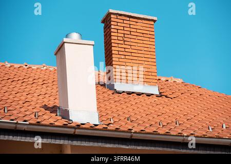 Zwei Schornsteine auf einem Ziegeldach mit Metallabdeckung und Regenrinnen gegen klaren blauen Himmel. Stockfoto