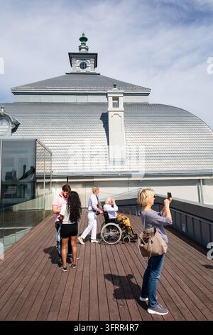 TOURISTEN AUF EINER DACHPLATFORM MIT AUSSICHTSTERRASSE, REISEBERICHT AUS RIGA, RIGA, LETTLAND: Das Lettische Nationalmuseum für Kunst. Eine Gruppe von Menschen, einige machen Fotos mit einem Mobiltelefon und ein anderer schiebt eine Person im Rollstuhl, steht auf einer hözernen Aussichtsplattform vor einem Gebäude mit einem markanten Dach. INFO: Riga, die Hauptstadt von Lettland, ist die größte Stadt der baltischen Staaten. Mit einer Mischung aus mittelalterlicher Geschichte und kosmopolitischem Charme ist es weltberühmt für seine atemberaubende, von der UNESCO gelistete Jugendstilarchitektur und eine wunderschön erhaltene Kopfsteinpflaster Altstadt. Stockfoto