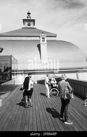 STÄDTISCHER SEHENSWERTEN AUF EINER AUSSICHTSPLATTFORM, RIGA REISEBERICHT, RIGA, LETTLAND: In einer Schwarzweißfotografie versammelt sich eine Gruppe von Menschen auf einem Holzdeck vor einem großen Gebäude, einschließlich einer Person im Rollstuhl und einer Frau, die mit ihrem Handy ein Foto macht. INFO: Riga, die Hauptstadt Lettlands, ist die größte Stadt der baltischen Staaten. Mit mittelalterlicher Geschichte und kosmopolitischem Charme vermischt, ist sie weltberühmt für ihre atemberaubende, von der UNESCO gelistete Jugendstilarchitektur und eine wunderschön erhaltene Kopfsteinpflaster Altstadt. Stockfoto