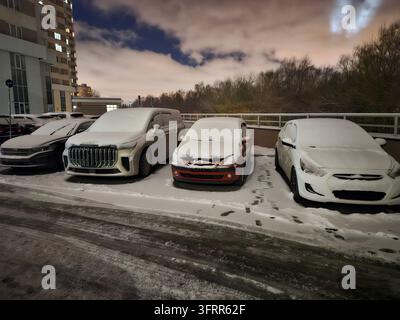 Vier Autos stehen auf einem Gästeparkplatz mit Schnee darauf. Die Autos sind ein rotes, weißes und schwarzes Auto, ein silbernes Auto und ein weißes Auto in einer Wohnanlage Stockfoto