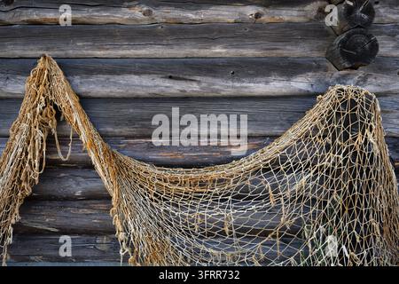 Eine Nahaufnahme eines alten, verwitterten Fischernetzes, das gegen eine rustikale Wand aus horizontalen Holzblöcken drapiert ist. Das dunkle, strukturierte Holz zeigt tiefe Kr Stockfoto