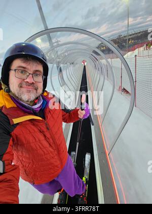 Mann mit roter Jacke und violetten Hosen steht in einem Skilifttunnel. Er hält einen Skistock und lächelt, macht Selfie Stockfoto