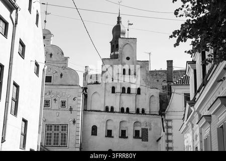 HISTORISCHE EUROPÄISCHE ARCHITEKTUR IN MONOCHROM, RIGA-REISEBERICHT, RIGA, LETTLAND: Die Drei Brüder (Trīs brāļi) ist der älteste Komplex von Wohnhäusern in Riga. Eine Schwarz-Weiß-Ansicht, die nach oben zu historischen Giebelgebäuden blickt, mit einem Kirchturm im Hintergrund. INFO: Riga, die Hauptstadt von Lettland, ist die größte Stadt der baltischen Staaten. Mit mittelalterlicher Geschichte und kosmopolitischem Charme vereint, ist sie weltberühmt für ihre atemberaubende UNESCO-geliste Art-Nouveau-Architektur und eine wunderschön erhaltene Kopfsteinpflaster-Altstadt. Stockfoto