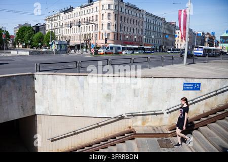 FRAU VERLÄSST EINE STADTUNTERFÜHRUNG INS SONNENLICHT, RIGA-REISEBERICHT, RIGA, LETTLAND: Eine junge Frau in einem schwarzen T-Shirt-Kleid steigt eine Betonstufentreppe hinauf, die aus einer Fußgängerunterführung hinausführt, in eine helle, sonnige Stadtstraße mit Verkehr und Gebäuden im Hintergrund. INFO: Riga, die Hauptstadt von Lettland, ist die größte Stadt der baltischen Staaten. Mit einer Mischung aus mittelalterlicher Geschichte und kosmopolitischem Charme ist sie weltweit bekannt für ihre atemberaubende UNESCO-gelistete Jugendstilarchitektur und eine wunderschön erhaltene Kopfsteinpflaster Altstadt. Stockfoto
