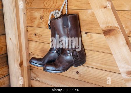 Braune Lederstiefel mit Gurtverzierungen, die an einem Seil gegen eine strukturierte Holzwand hängen, und einen rustikalen und Vintage-Stil evozieren Stockfoto