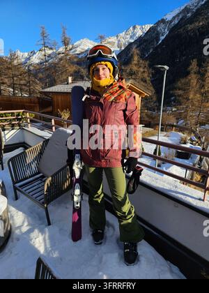Eine Frau in einer roten Jacke und einer grünen Hose steht mit einem Skistock in der Hand auf einem Balkon in einem Skiressort Stockfoto