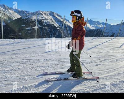 Eine junge Frau in einer roten Jacke und einer Schutzbrille steht auf Skiern auf einem schneebedeckten Hang in einem Skiresort nahe eines Zauns Stockfoto