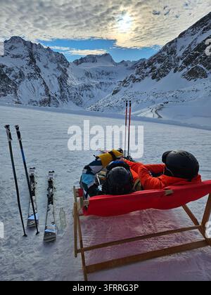 Ein Paar Skifahrer entspannt sich auf einem roten Stuhl im Schnee. Der Stuhl steht auf einer Holzplattform. Die Skifahrer tragen Skier und Helme. Die Szene ist friedlich und ruhig im Skiresort Stockfoto