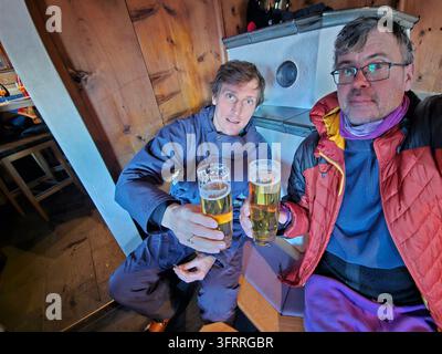 Zwei Männer sitzen nebeneinander und halten ihre Biergläser hoch. Sie lächeln und genießen ihre Getränke in der Nähe des Ofens im Skiresort Stockfoto