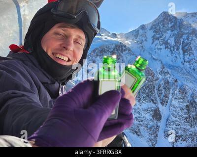 Ein Mann hält zwei Flaschen Alkoholdrink in den Händen mit einem Freund und lächelt. Die Flaschen sind grün und der Mann trägt eine schwarze Jacke auf dem Skiresort Stockfoto