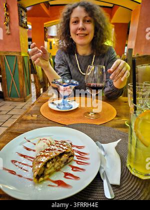 Moskau - 12. Feb. 2025: Eine Frau (MR) sitzt an einem Tisch mit einem Dessert und einem Glas Wein. Sie lächelt und genießt ihre Mahlzeit in mexikanisch Stockfoto