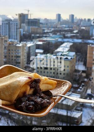 Ein köstlicher Dessertpfannkuchen mit Marmelade auf Maslenitsa wird auf einem kleinen Teller serviert, mit einem Löffel darauf, Blick auf die Skyline der Stadt im Hintergrund, wodurch eine gemütliche und einladende Atmosphäre entsteht Stockfoto