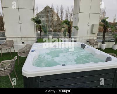 Moskau - 10. März 2025: Ein großer, weißer Whirlpool steht auf einem grasbewachsenen Dachgarten. Der Whirlpool ist von mehreren Stühlen umgeben, und im Hintergrund ist eine Palme zu sehen. Konzept von Entspannung und Freizeit Stockfoto