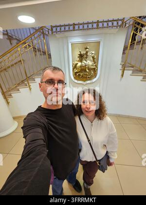 MINSK - 27. MÄRZ 2025: Ein Mann und eine Frau (MR) posieren für ein Foto in einem Gebäude mit einem goldenen Statuengrundriss des Wappens von Moskau. Der Mann trägt ein schwarzes Hemd und eine Brille Stockfoto