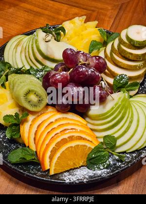 Ein Teller Obst mit einer Vielzahl von Äpfeln, Orangen und Trauben. Der Teller mit Birne, Kiwi steht auf einem Holztisch Stockfoto