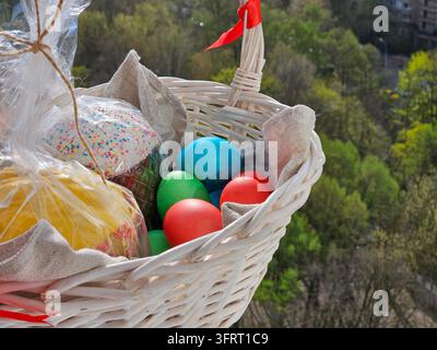 Korb mit Ostereiern und ein Kuchen. Der Korb ist weiß und hat ein rotes Band. Die Eier sind in verschiedenen Farben und Größen Stockfoto