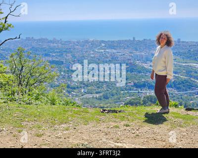 Frau steht auf einem Hügel mit Blick auf eine Stadt. Sie trägt eine weiße Jacke. Der Hügel ist mit Gras bedeckt und im Hintergrund stehen Bäume Stockfoto