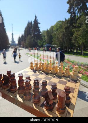 Ein hölzernes Schachbrett mit schwarzen und weißen Figuren wird von jemandem hochgehalten. Das Brett steht auf einem Bürgersteig vor einem Flussuferbahnhofspark. Die Szene ist friedlich und ruhig Stockfoto