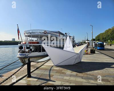 Moskau – 26. Juli 2025: Ein Boot liegt neben einem weißen Papierboot festgemacht. Das Boot ist ein großes Schiff mit einer roten Flagge darauf. Die Szene ist friedlich und ruhig, mit dem Wasser im Flussuferbahnhof aus der Stalin-Ära Stockfoto