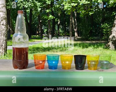 Eine Flasche Bier oder Kvass wird in sechs bunte Plastikbecher gegossen. Die Becher sind in einer Reihe auf einem Tisch angeordnet, wobei die Bierflasche in der Mitte steht. Die Szene vermittelt eine lässige und entspannte Atmosphäre Stockfoto