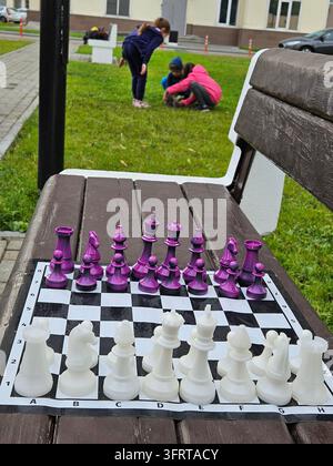 Ein lila und weißes Schachset liegt auf einem Holztisch. Eine Gruppe von Kindern spielt auf dem Gras hinter dem Tisch Stockfoto