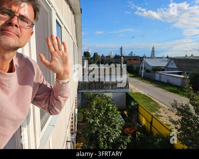 Mann steht auf einem Balkon und winkt jemandem zu. Die Szene spielt sich in einem Wohngebiet mit Häusern und Bäumen im Hintergrund ab Stockfoto