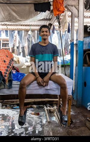 Porträt eines lächelnden jungen Mannes, der auf einem Tisch in einer Wäscherei sitzt, mit Jeans, die zum Trocknen an Dhobi Ghat, Maharashtra, Mumbai, Indien hängen Stockfoto