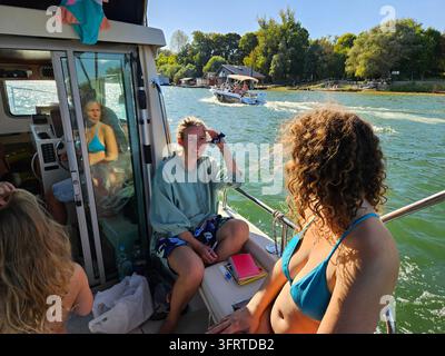 Gruppe von Menschen befindet sich auf einem Boot, wobei eine Frau auf dem Heck des Bootes sitzt und eine andere Frau auf der Vorderseite sitzt. Die Frau auf dem Heck trägt einen Bikini und lächelt Stockfoto