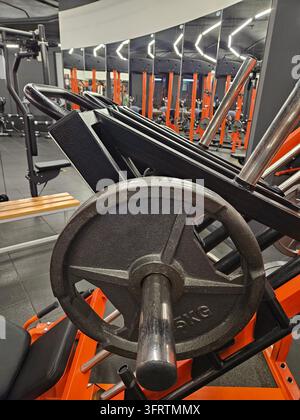 Gewichtsteller ruht auf Fitnessgerät. Orangener Rahmen hält schwarze Metallstange. Spiegelfliesen spiegeln Fitnessgeräte wider. Stahl-Hantelstange liegt untätig neben der Scheibe. Beleuchtung leuchtet über dem Fitnessraum Stockfoto