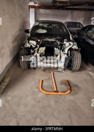 Beschädigtes Auto steht in einem Beton-Garagenraum. Eine orangefarbene Absperrung ruht auf der lackierten Bodennummer. Motor und Rahmen des Fahrzeugs sind freigelegt. Garagenwände und Decke Stockfoto