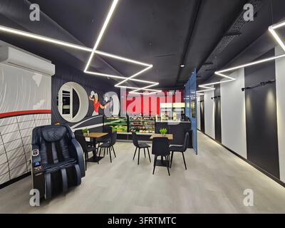 Moskau - 08. Okt. 2025: Moderner Bürolounge mit schwarzen Deckenleuchten und Stühlen. Ein blauer Massagestuhl steht neben einem weißen Wandgemälde. Rote Akzentwand mit großem kreisförmigem Design und Regalen Stockfoto