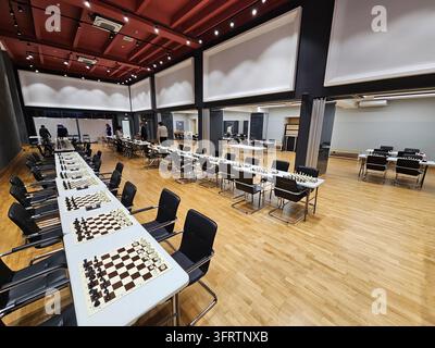 Moskau – 6. Dez. 2025: Moderne Schachhalle mit langen Tischen und Stühlen. Weißtafeln für das Turnierspiel aufgestellt. Holzböden und rot akzentuierte Decke. Stockfoto