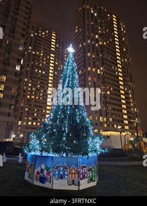 Weihnachtsbaum leuchtet nachts hell. Der Baum steht auf einem festlich geschmückten Sockel. Hohe Gebäude leuchten dahinter. Fenster leuchten warm gegen die Dunkelheit Stockfoto