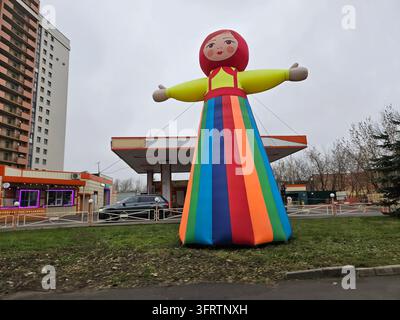 Moskau - 06. Dez. 2025: Große aufblasbare Puppe steht in der Nähe einer Tankstelle. Helle Farben schmücken ihr langes, gestreiftes Kleid. Ein kindliches Gesicht zeigt rotes Haar und weite Augen. Ausgestreckte Arme rahmen die Puppe ein Stockfoto