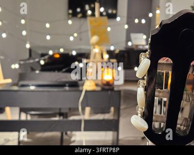 Nahaufnahme des Gitarrenkopfes mit weißen Stimmwirbeln. Sanft beleuchteter Raum mit Lichterketten im Hintergrund. Klavier sitzt dahinter, leicht unscharf. Warmes Lampenlicht verleiht eine gemütliche Atmosphäre Stockfoto