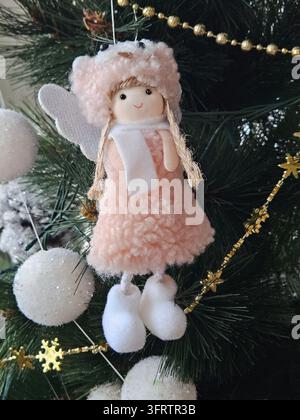 Weiche rosa Engelspuppe hängt am Weihnachtsbaum. Flauschiges Kleid und Flügel verleihen sanften Charme. Weißer Schal und Stiefel vervollständigen das Aussehen. Goldene Perlen und Schneebälle schmücken den Baum Stockfoto