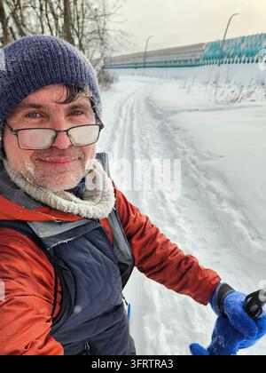 Mann lächelt in einer verschneiten Winterlandschaft. Mit Brille, Hut und Handschuhen steht er im Schnee. Der Weg hinter ihm zeigt Spuren und einen fernen Zaun. Schnee Stockfoto