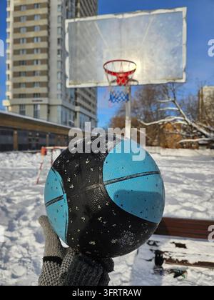 Hand hält einen blauen und schwarzen Basketball im Schnee. Ein Basketballkorb steht hinter schneebedecktem Boden. Die helle Sonne scheint durch den klaren Winterhimmel. Schneeiger Stadtpark mit einem hohen Gebäude in der Nähe Stockfoto